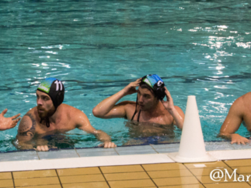 Un grupo de jugadores de waterpolo conversando en la piscina, todos con cascos y ropa deportiva. El agua es clara y brillante, reflejando la luz del día. La pared de la piscina está decorada con azulejos amarillos y verdes.