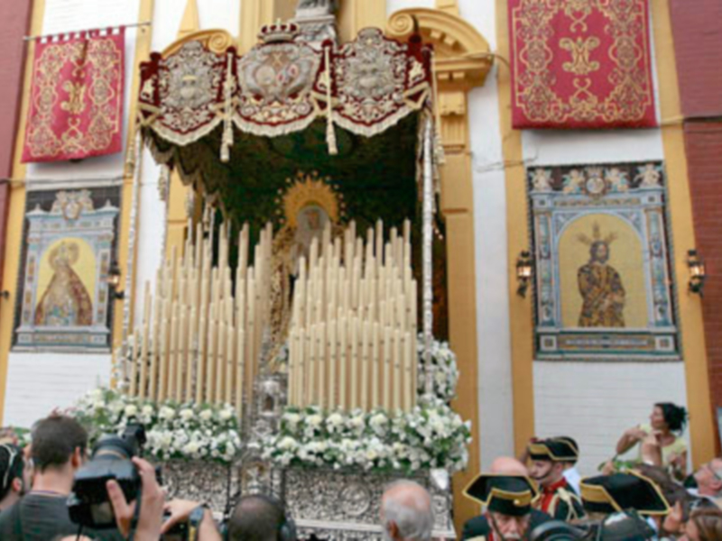 Fotografía de una procesión religiosa con un altar central adornado con velas y flores, rodeado por personas en trajes históricos. En el fondo, se observan carteles y una estructura arquitectónica con detalles dorados. La imagen transmite la solemnidad y el fervor de una celebración religiosa tradicional.