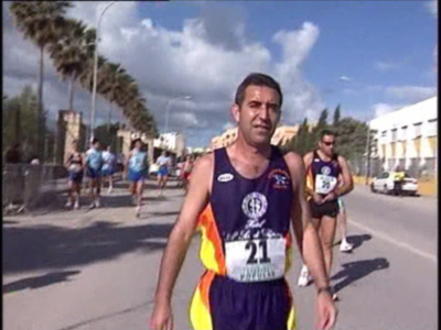 Maratón en una carretera con palmeras y edificios a ambos lados. El atleta en primer plano lleva un número 21 y una camiseta con el símbolo de la UE. Otros corredores están en segundo plano, también con números y camisetas distintas. El cielo es azul con algunas nubes.