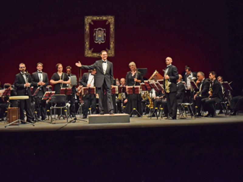 Orquesta en escenario con músicos tomando notas y un director al frente