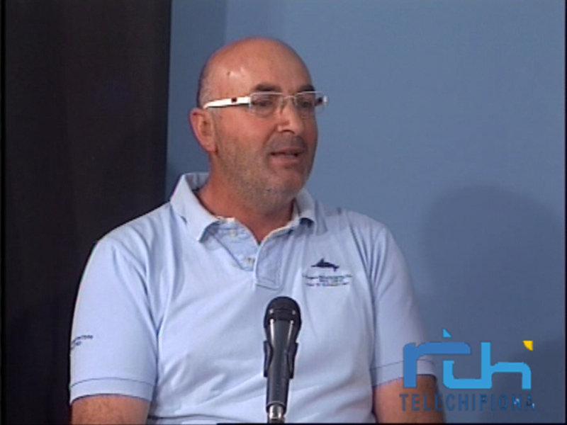 Un hombre con gafas habla en un evento público, posiblemente en un evento deportivo o de negocios, con un fondo azul y una insignia en su camisa.