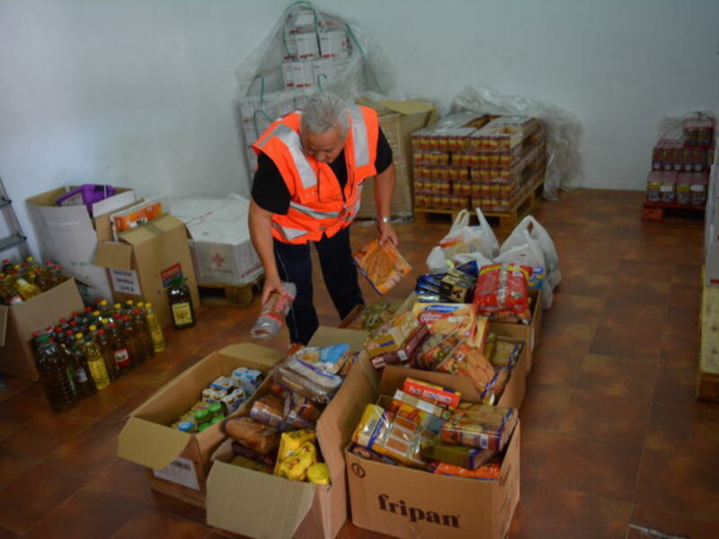 Un voluntario prepara paquetes de alimentos en una estantería de un almacén, rodeado de cajas y paquetes de productos de primera necesidad.