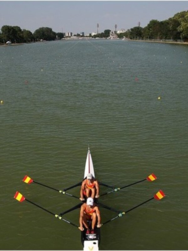 Dos personas remando en un bote de remo en el río, con árboles y cielo azul en el fondo.