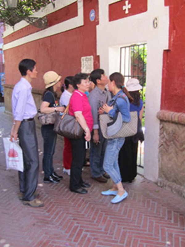Una fila de personas esperando en una calle con tiendas y un edificio rojo con una cruz roja.