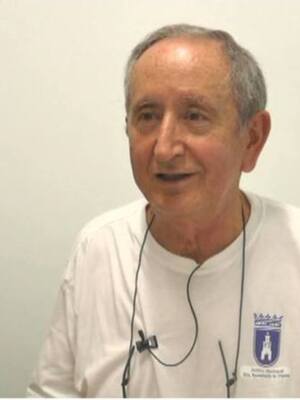 El hombre está sonriendo y parece estar en una entrevista o charla. La imagen es clara, con un fondo blanco y el hombre viste una camiseta blanca con detalles azules.