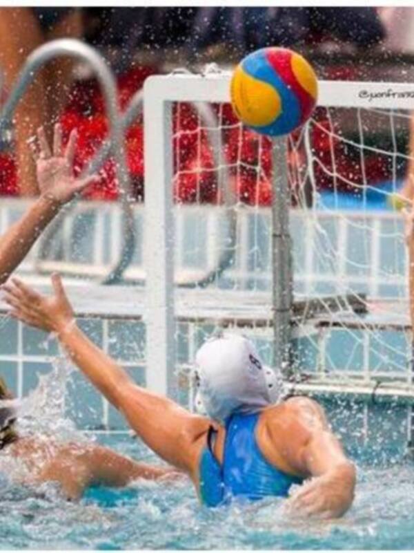 Un equipo de waterpolo compite en un partido. Los jugadores se mueven rápidamente, lanzando y bloqueando la pelota de colores. La portería está claramente visible en el fondo, con un equipo rival esperando su turno. La acción es intensa y dinámica en el agua azul.