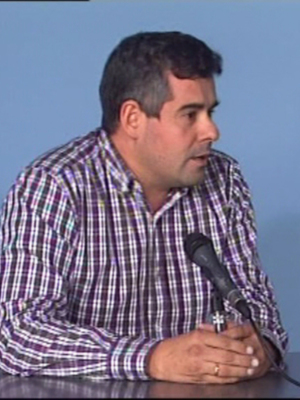 Un hombre con una camisa a cuadros habla en un micrófono durante una conferencia. La imagen muestra su rostro y parte de su torso, con un fondo azul claro.
