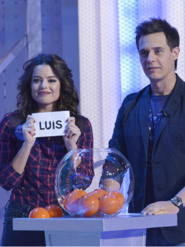 Un hombre y una mujer están en un escenario con una pantalla de fondo. La mujer sostiene una tarjeta con la palabra "LUIS" y sonríe mientras el hombre, vestido con una chaqueta negra, observa. En la mesa delante de ellos hay frutas y un bol transparente con una figura en su interior.