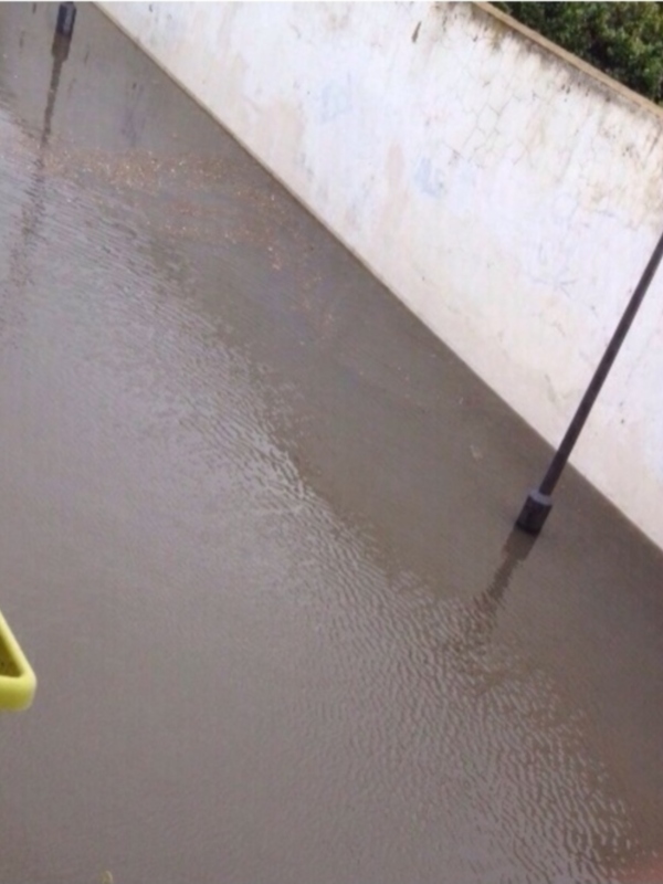 Fotografía de una calle inundada con agua hasta la mitad del asfalto, cerca de un muro blanco y una lámpara de calle. Un contenedor amarillo está parcialmente cubierto por el agua.