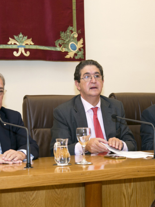 Tres hombres en trajes sentados en una mesa con micros y copas de vino, frente a una bandera decorada.
