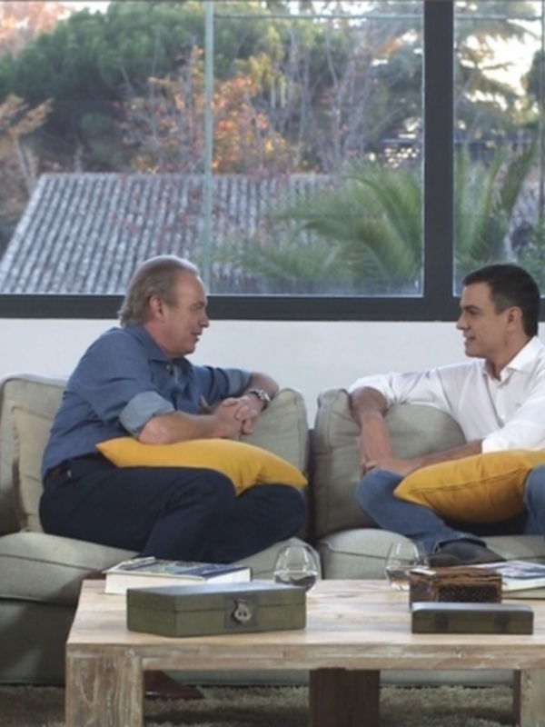 Dos hombres conversan en un sofá beige con cojines amarillos, frente a una ventana que revela un paisaje con árboles y una casa en el horizonte.