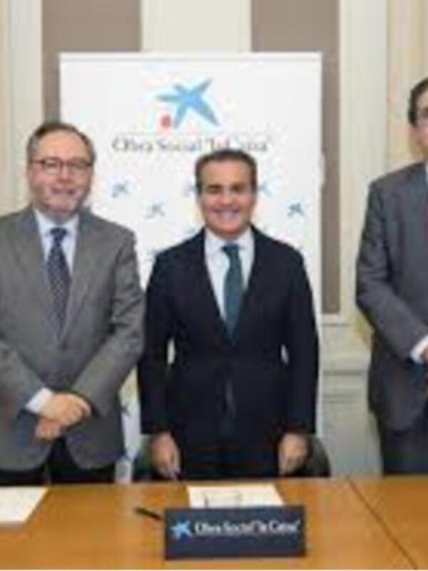 Tres hombres en trajes estrechando manos en una sala de conferencias con un logo en el fondo.