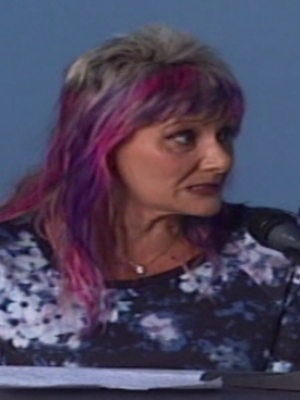Una mujer con cabello rosa y azul está hablando en un micrófono frente a una pantalla de fondo azul.