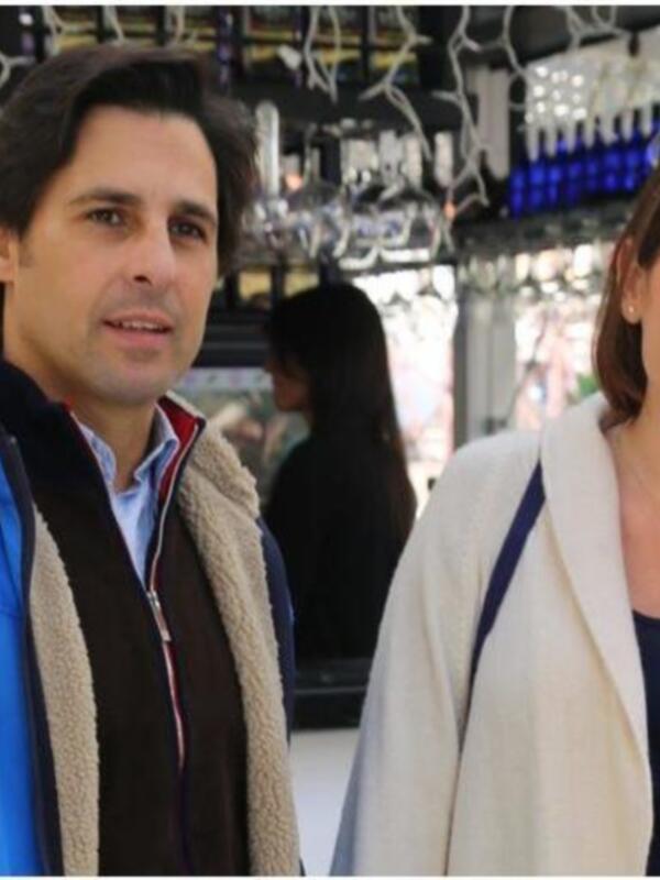 Dos personas posan frente a una barra de botellas. La persona izquierda lleva una chaqueta azul con capucha beige, mientras que la derecha usa un abrigo blanco. Ambos tienen cabello largo y sonríen. La barra de botellas está en el fondo, con varios tipos de bebidas.