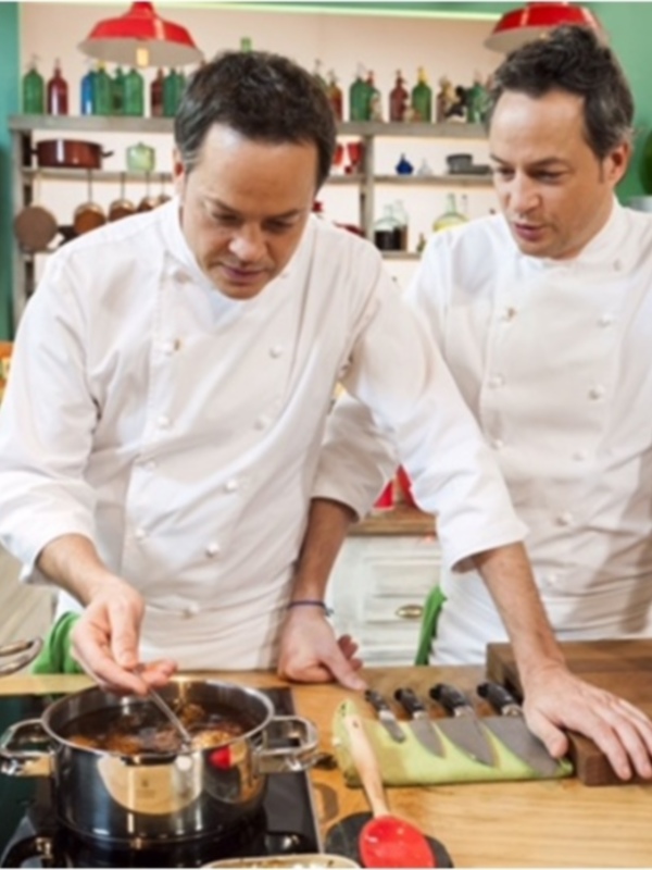 Dos chefs en una cocina profesional, uno cocinando en una olla y el otro observando, con un fondo de estanterías con vinos y utensilios de cocina.