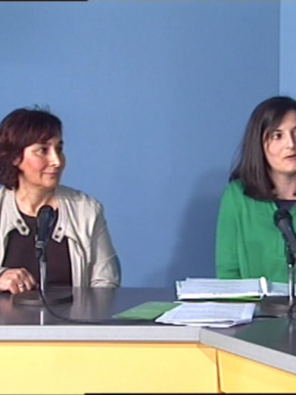 Dos mujeres sentadas en una mesa frente a un fondo azul, con micrófonos y documentos en el frente.