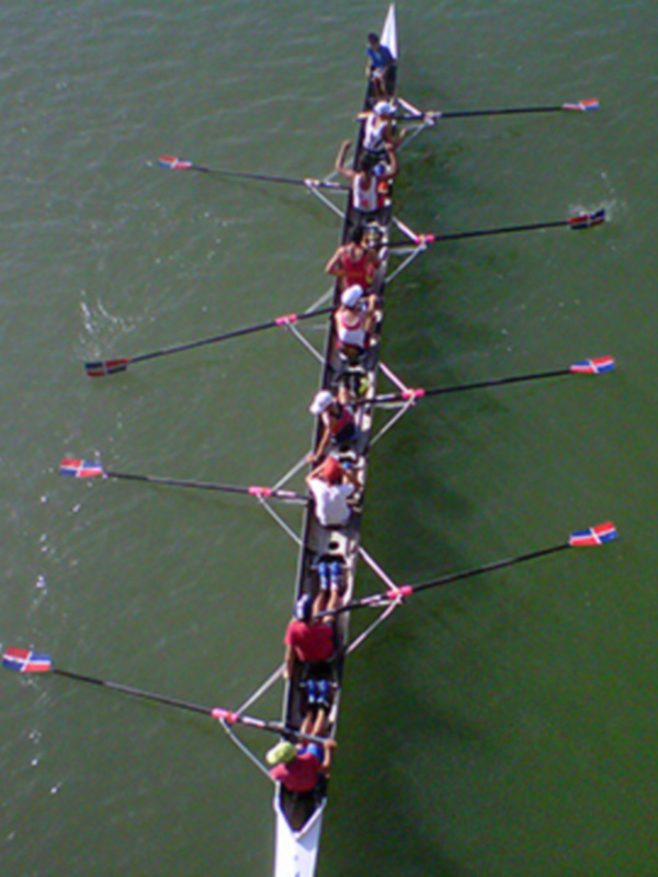 Equipo de remo en acción en el agua, con remos levantados y banderas rojas, azules y blancas en la punta de los remos.