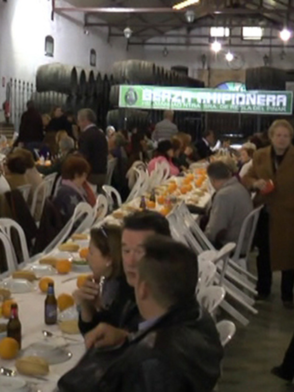 Una multitud de personas disfruta de una comida en un ambiente acogedor con mesas llenas y botellas de cerveza. La decoración incluye estantes con barriles y una gran pantalla que anuncia "Pizzeria Panadería". La atmósfera es festiva y alegre.