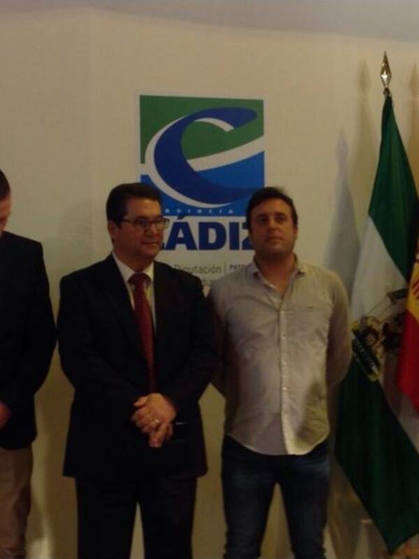 Presentación de un evento en el que cuatro hombres, dos con trajes y dos con camisas, se encuentran frente a una pantalla de televisión azul y blanca. En el fondo, hay banderas españolas y una bandera de Andalucía.
