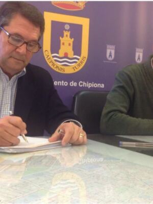 Dos hombres firman un documento en una mesa con un mapa de Chipiona al fondo.