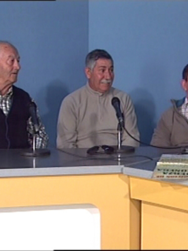 Tres hombres sentados en una mesa de conferencia, con micrófonos y un libro abierto. El ambiente es formal y profesional.