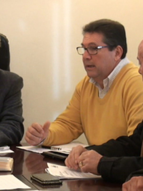 Grupo de personas sentadas en una reunión, probablemente un consejo o comité. La imagen muestra a cuatro personas sentadas alrededor de una mesa con documentos y teléfonos. La escena sugiere un ambiente formal o profesional, posiblemente una reunión de trabajo o planificación.