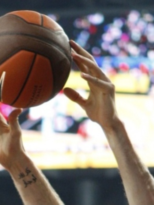 Una persona sostiene un balón de baloncesto en un escenario de competición, con el anillo de la canasta visible en el fondo.