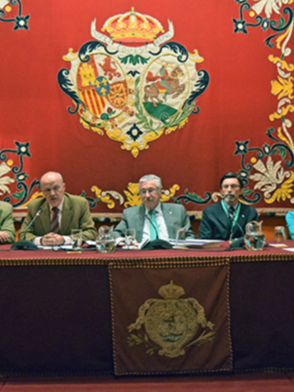 Conferencia en un salón con escudo real español en el fondo, participantes sentados en una mesa con documentos y vasos de cristal.