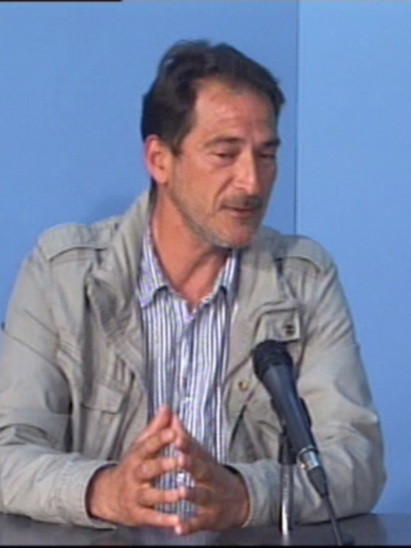 Un hombre hablando en una conferencia, con un fondo azul claro y una silla azul en el fondo.