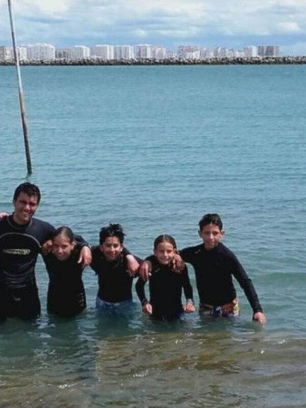 Una familia en trajes de neopreno posando en el agua cerca del mar, con rocas y edificios al fondo.