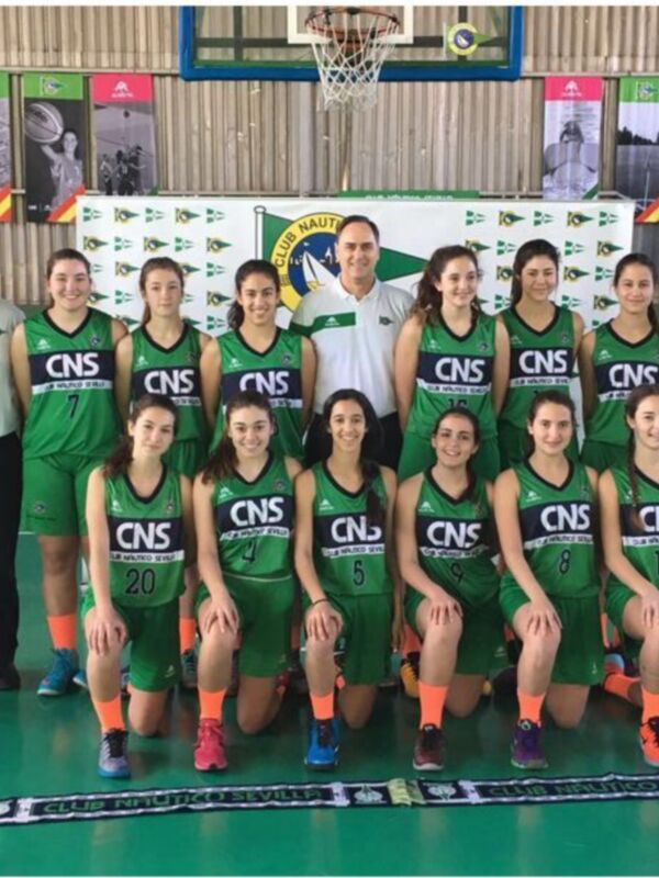 Equipo femenino de baloncesto posando en el estadio del Club Nautico de Sevilla, con camisetas del equipo y una bandera del club en el fondo.
