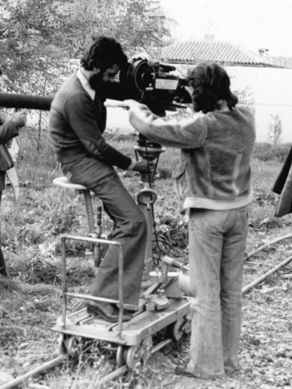 Directores de cine en el set de rodaje, con una cámara en primer plano y otros personajes en el fondo, en una escena de producción cinematográfica.