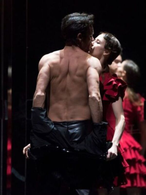 Un hombre sin camisa y una mujer en un vestido rojo se besan en una escena dramática.