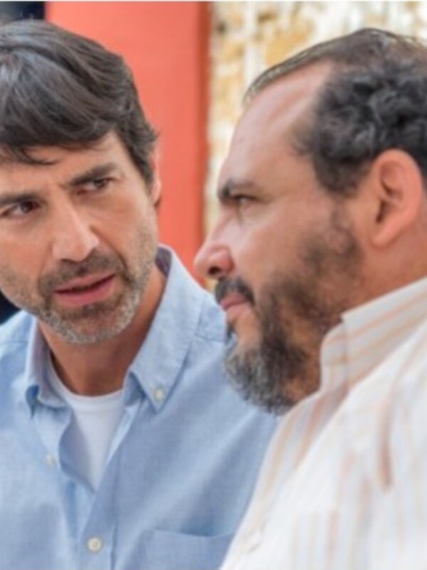 Dos hombres conversan en un ambiente informal, uno con camisa azul y el otro con una camisa de rayas. La escena sugiere un diálogo serio o profundo entre ellos.