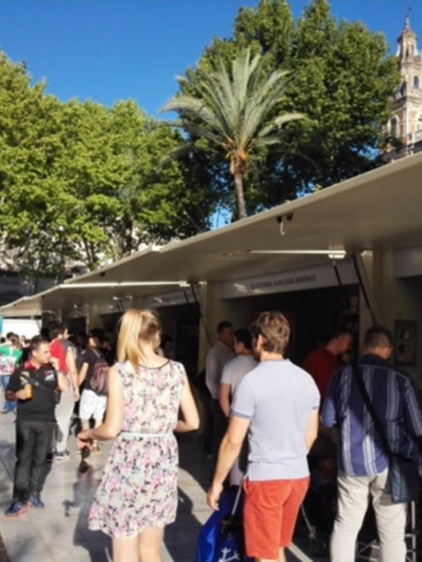 Un grupo de personas caminando por un mercado al aire libre en un día soleado, con árboles y una torre en el fondo.