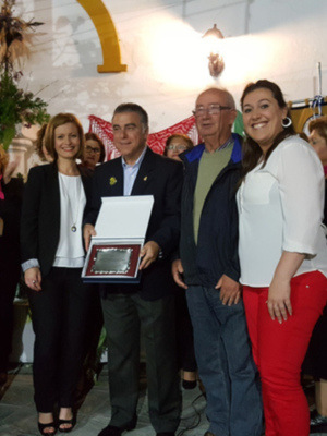 Una imagen de una celebración festiva con personas sonriendo y posando para la foto. En el centro, una persona sostiene un reconocimiento mientras otros asistentes observan con alegría. La decoración incluye banderas y flores, creando un ambiente festivo.