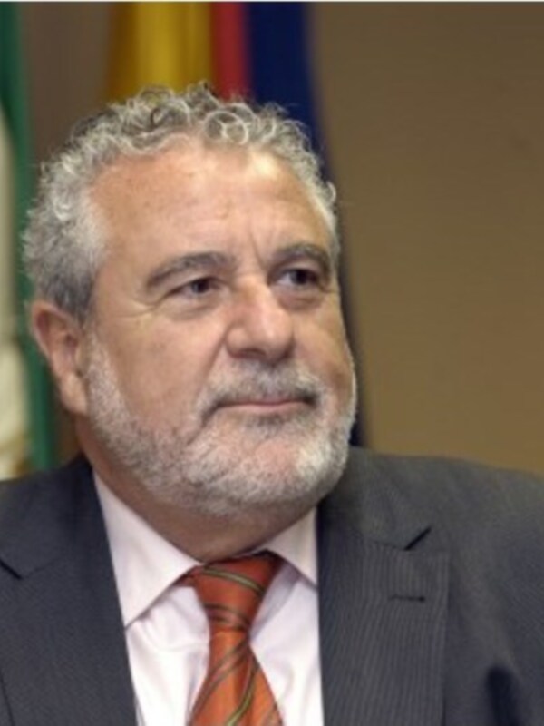 Un hombre con cabello canoso y barba gris, vistiendo una camisa blanca y corbata naranja. Está sentado en un entorno formal con una bandera en el fondo.