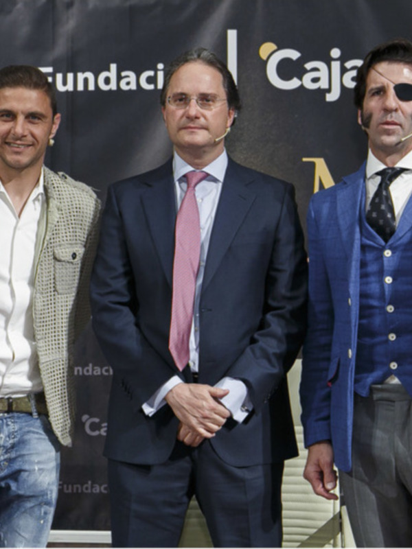 Tres hombres posan frente a un fondo con el logo de Cajasol y Fundación. El hombre en medio lleva una chaqueta beige, el de la izquierda una camisa blanca y vaqueros, mientras que el hombre en la derecha lleva un traje azul con una camisa blanca y corbata.