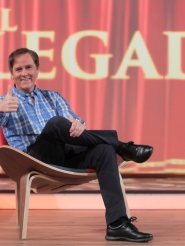 Un hombre está sentado en un sillón de madera con una camisa azul y pantalones negros, sonriendo y mostrando un gesto de aprobación con su mano derecha. El fondo es rojo con letras doradas que dicen "EL DELEGADO".