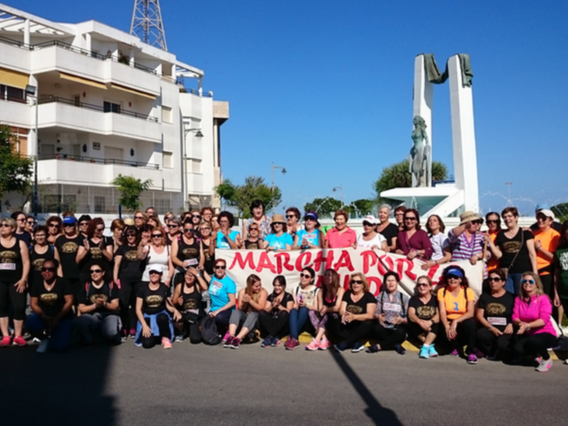 Una multitud de personas posa en una calle con un cartel que dice "MATUHA PON". En el fondo, se puede ver una estructura blanca y un monumento con una figura en alto. La escena se desarrolla bajo un cielo azul claro, y a la derecha hay una calle con algunos vehículos.