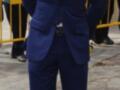 Un hombre de pie en una calle, vistiendo un traje azul con detalles en negro y un corbata roja. Detrás de él, hay una valla amarilla y otros individuos que parecen estar en un evento formal.