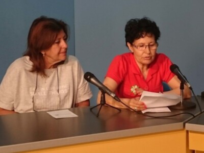 Dos personas sentadas detrás de una mesa con micrófonos, sosteniendo papeles y discutiendo.