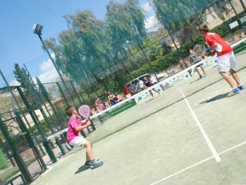Niños jugando tenis en una cancha de tenis al aire libre, con un público observando desde el lado.