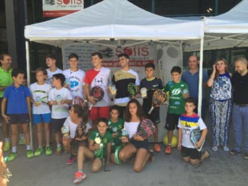 Un grupo de niños y adultos posan bajo un toldo en una feria o evento, con camisetas de fútbol y pelotas. El ambiente es festivo y familiar, con una diversidad de edades representadas.