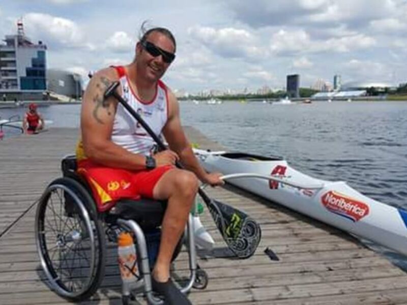 Un atleta paralímpico en silla de ruedas posa con su canoa en el río, con el estadio olímpico al fondo.
