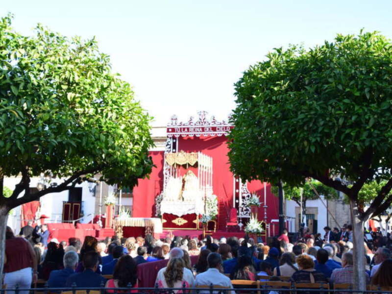 Festividad religiosa en un pueblo con una imagen de la Virgen en el centro, rodeada de árboles y una multitud de personas observando la procesión.