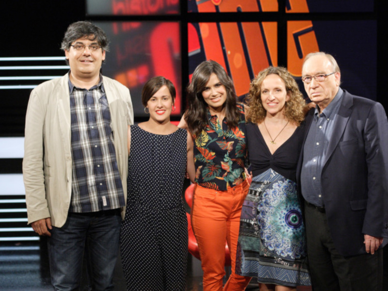Una foto de grupo de cuatro personas en una escena de teatro o televisión, con una mujer en el centro y tres personas a su izquierda, una mujer en el centro y dos hombres a su derecha. El fondo es oscuro con luces rojas y blancas.
