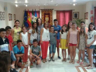 Grup de nens i nenes posant a l'interior d'un edifici. Al fons, es poden veure banderes i un escut d'armes. Els nens i nenes estan vestits amb ropa de colors vivos, amb pantalons roges i blaus. La imatge està il·luminada amb llum natural, probablement de la finestra.