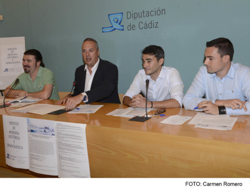 Sala de prensa de la Diputación de Cádiz con cuatro personas sentadas en una mesa. En el fondo, se puede ver un cartel con la bandera de Cádiz y el nombre de la institución. La mesa tiene documentos en español, lo que sugiere una reunión o conferencia de prensa.