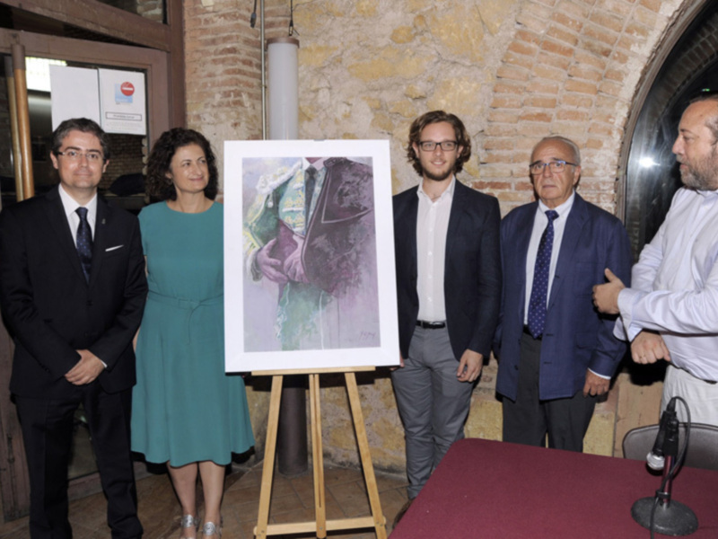 Un grupo de personas posa frente a un lienzo con una obra de arte en el interior de un edificio histórico. La imagen muestra a cuatro personas, tres hombres y una mujer, vestidos formalmente. En el fondo se puede ver una pared de ladrillo y un cartel con información sobre la exposición. La escena sugiere una presentación o evento artístico en un lugar de importancia cultural.
